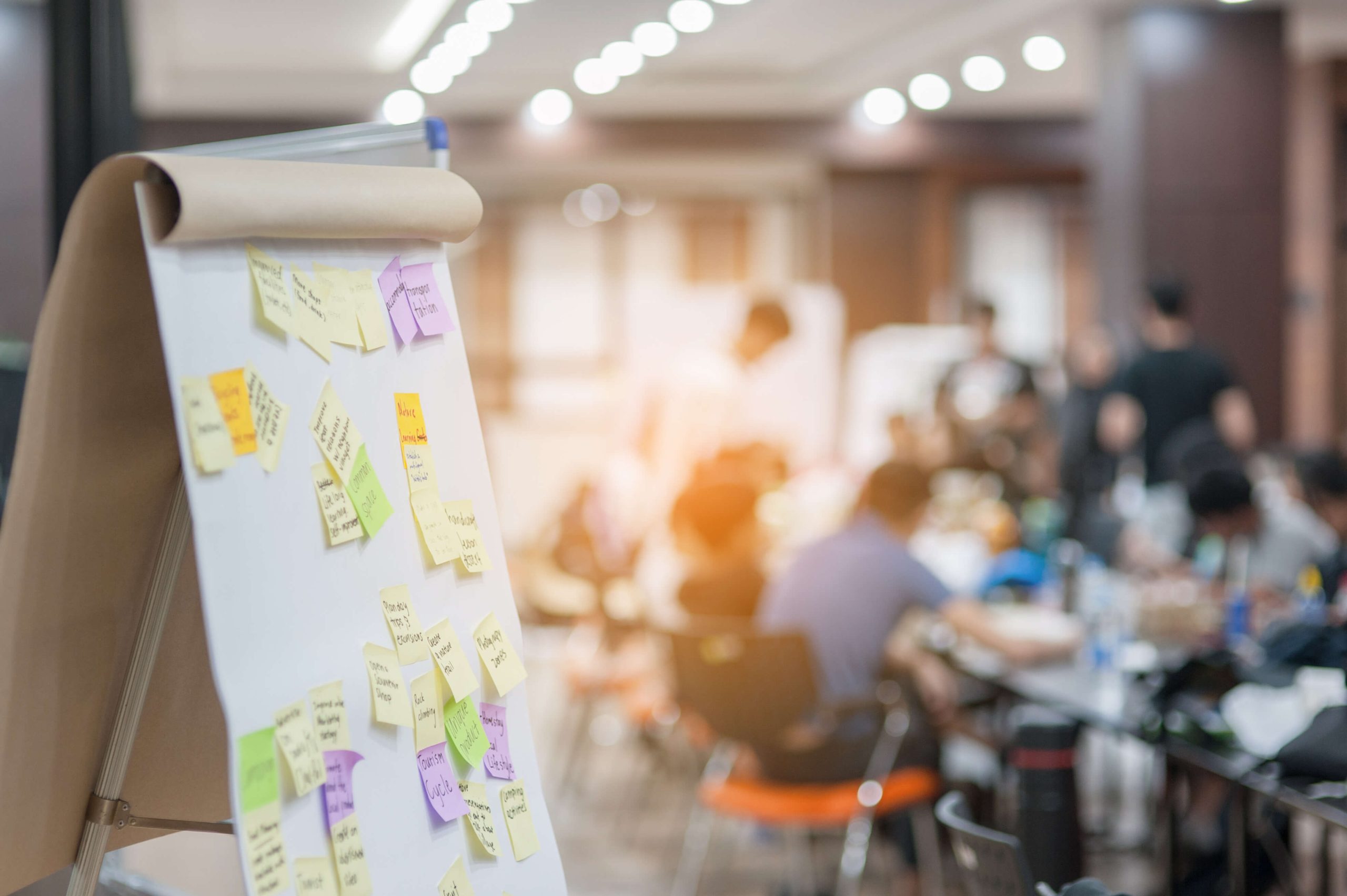 Mehrere Personen sitzen in einem Seminarraum und beraten sich, im Vordergrund ist ein Whiteboard mit Post its zu sehen.